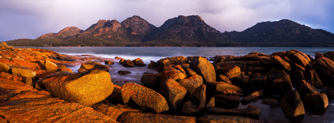Freycinet Coast by Mitchell Burns — warm sunlight on orange granite boulders, blue water and the Hazards range in Tasmania — illustrative Black + Black corner detail